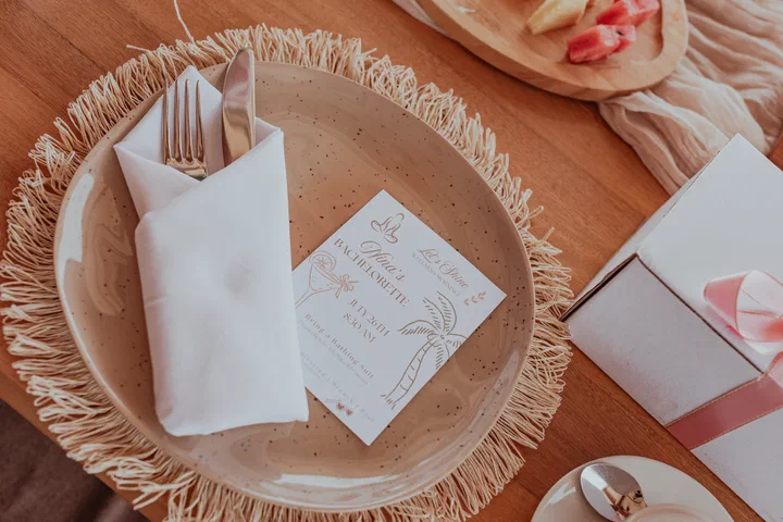 A place setting seen from above, with a speckled plate on a woven charger, a folded napkin with cutlery and a printed menu card.