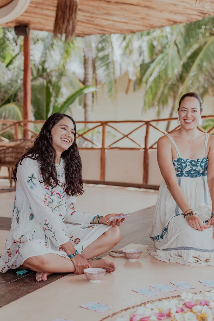Two friends laugh as they sit on the floor during a ceremony on a terrace surrounded by palms.