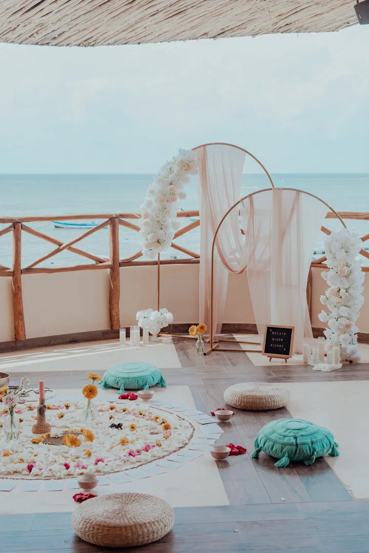 A seaside terrace set for a ceremony, with a flower mandala on the floor, cushions and draped arches with white flowers.