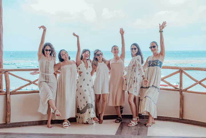 A group of friends in flowing dresses raise their arms on a terrace above the sea.