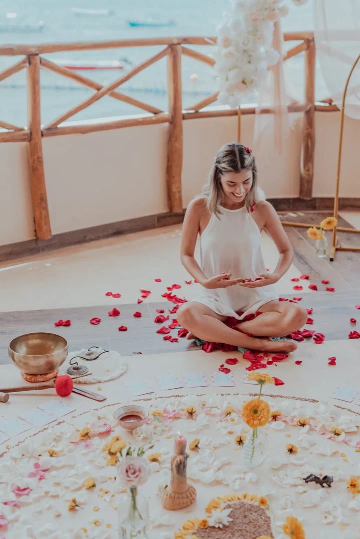 A woman sits cross-legged among scattered rose petals beside a flower mandala and a singing bowl on a seaside terrace.
