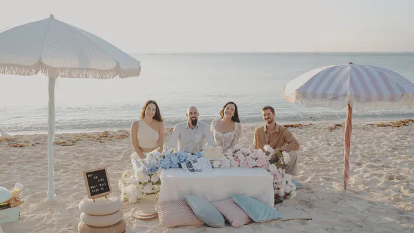 Four guests seated at a blue-and-pink picnic setup on the beach at sunrise