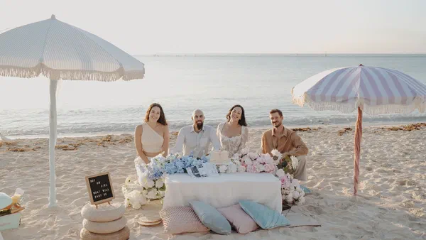 Four guests seated at a blue-and-pink picnic setup on the beach at sunrise