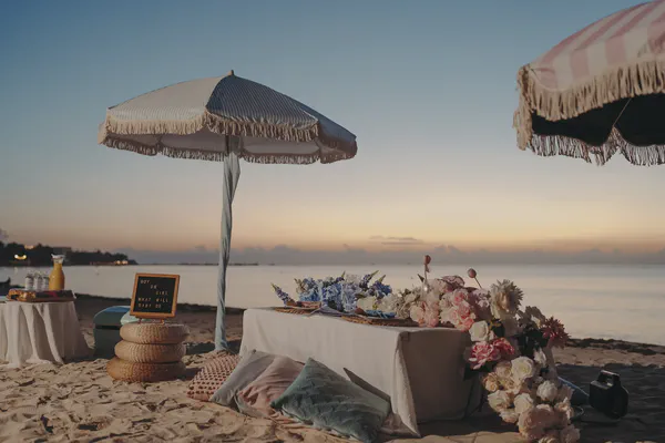 Sunrise beach picnic decor with striped umbrellas, blue and pink flowers, pillows, and a low table