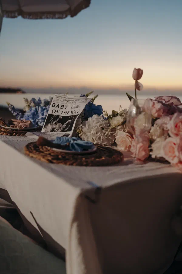 Blue and pink beach picnic table with flowers, woven place settings, cake, and a baby announcement newspaper