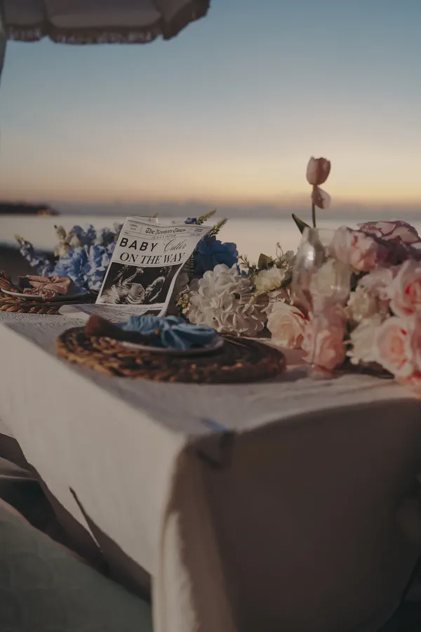 Blue and pink beach picnic table with flowers, woven place settings, cake, and a baby announcement newspaper
