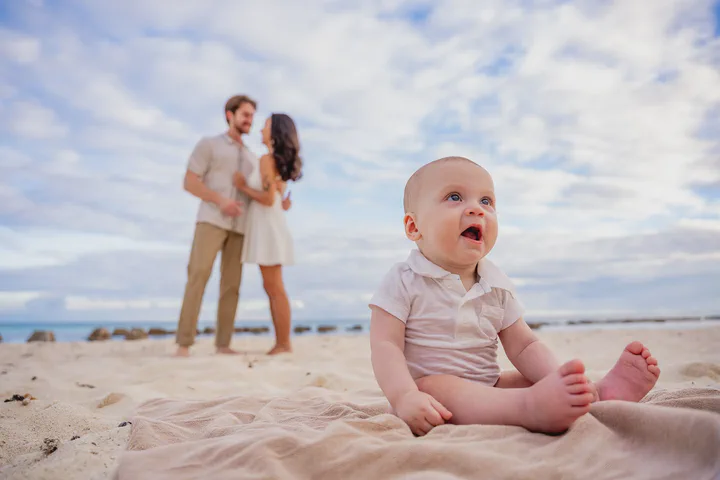 Baby sitting on the sand with smiling parents in the background