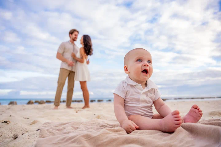 Baby sitting on the sand with smiling parents in the background