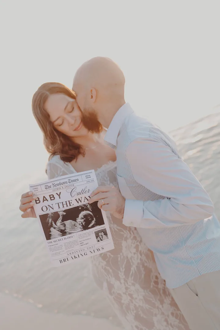 Expectant couple holding a Baby on the Way newspaper on the beach