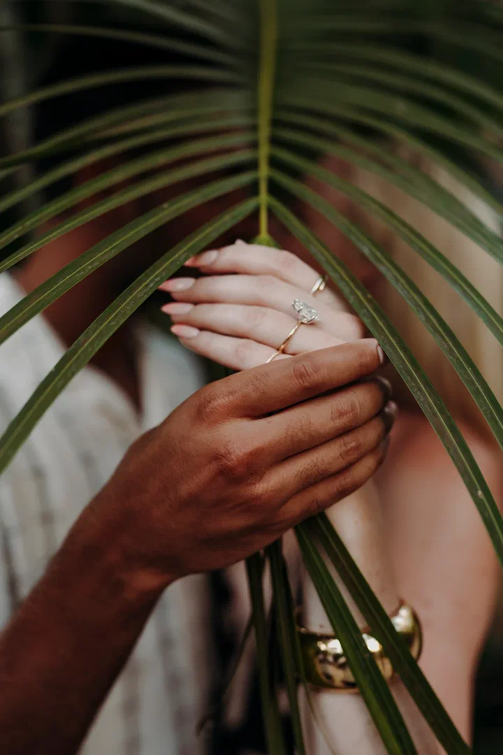 Close-up of a couple’s hands and an engagement ring behind a palm leaf