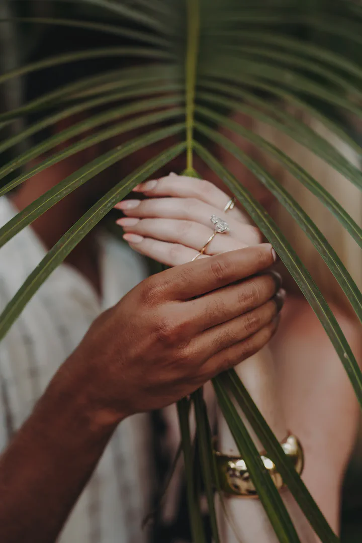 Close-up of a couple’s hands and an engagement ring behind a palm leaf