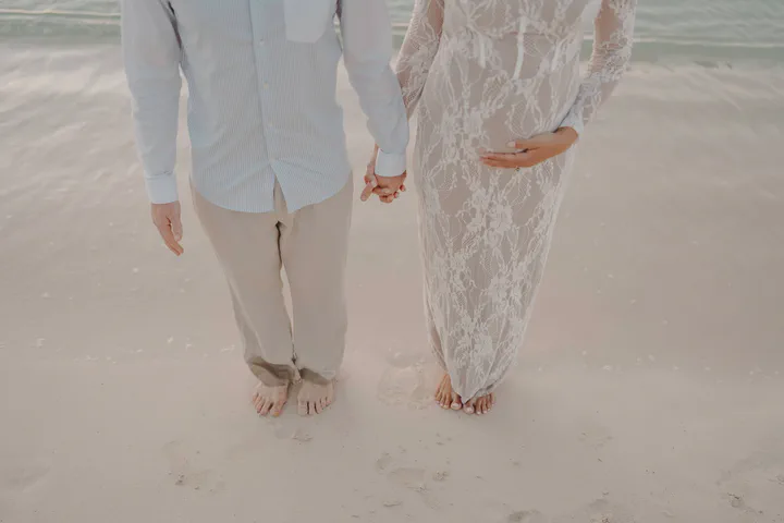 Expectant couple holding hands and walking barefoot on pale sand
