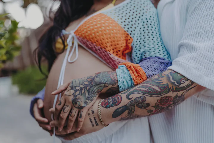 Tattooed hands resting on an expectant mother’s belly beneath a colorful crochet top