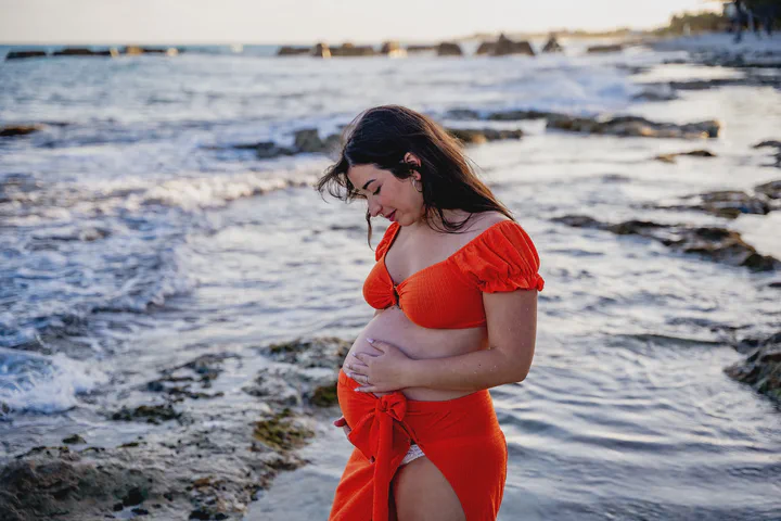 Expectant mother in a flowing red dress beside waves and coastal rocks