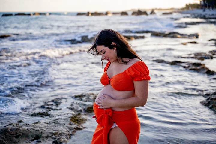 Expectant mother in a flowing red dress beside waves and coastal rocks