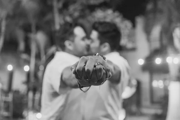 Fotografía en blanco y negro que capta de forma íntima manos entrelazadas con anillos de compromiso, sobre un fondo suavemente difuminado con luces de cadena; la composición evoca elegancia atemporal y un ambiente romántico con sutil toque tropical.