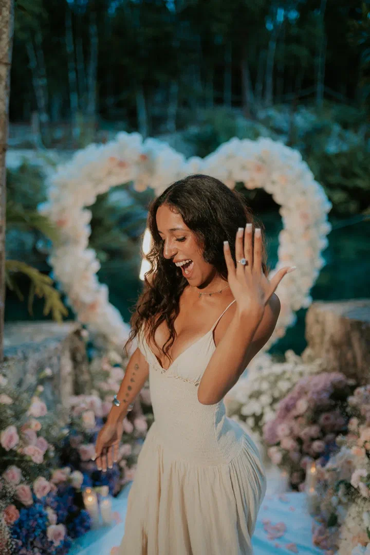 Una mujer muestra su anillo de compromiso frente a un corazón de flores blancas en la selva
