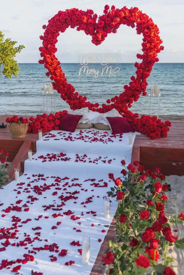 Propuesta de matrimonio en Playa Pescadores de Tulum con un magnífico arco en forma de corazón elaborado con rosas rojas vibrantes; un corredor blanco adornado con pétalos y un letrero de neón se integran con portavelas de vidrio y cojines burgundy.