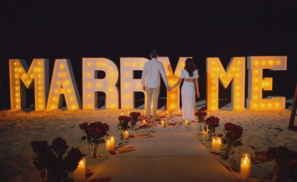 Impresionante propuesta nocturna en la playa, donde grandes letras iluminadas 'MARRY ME' irradian un resplandor dorado sobre arena inmaculada; un camino flanqueado por velas en portavelas de cristal y ramos de rosas rojas intensas crea un ambiente dramático y romántico.