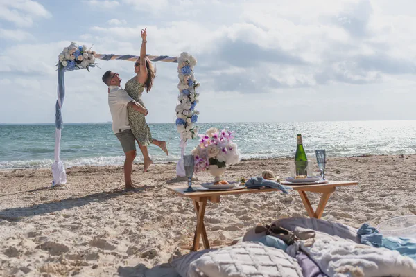 Celebración costera vibrante en Playa Xpu-Ha con un elegante arco floral adornado con rosas blancas y azules, enmarcado por un impresionante océano turquesa; se acompaña de un lujoso picnic con mesa de madera, cristalería fina y champagne.