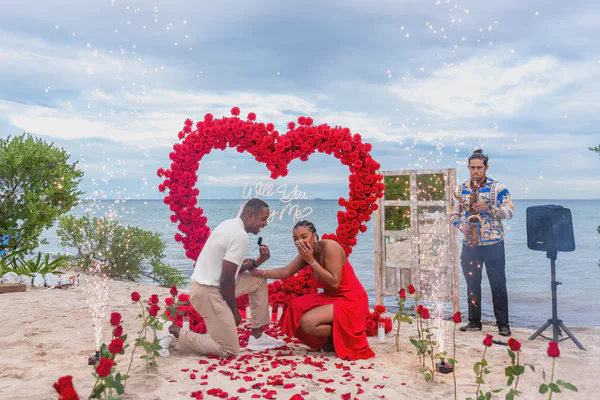 Propuesta de matrimonio en la icónica Playa Paraíso de Tulum con un imponente arco en forma de corazón elaborado con rosas rojas vibrantes; un letrero de neón 'Will You Marry Me?' y fuentes de chispas frías iluminan la escena sobre arena beige, realzando el equilibrio perfecto entre lujo y belleza natural.