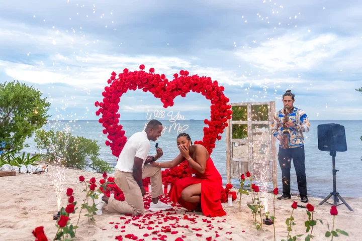 Hombre arrodillado pidiendo matrimonio frente a un corazón de rosas rojas en una playa del Caribe mientras toca un saxofonista