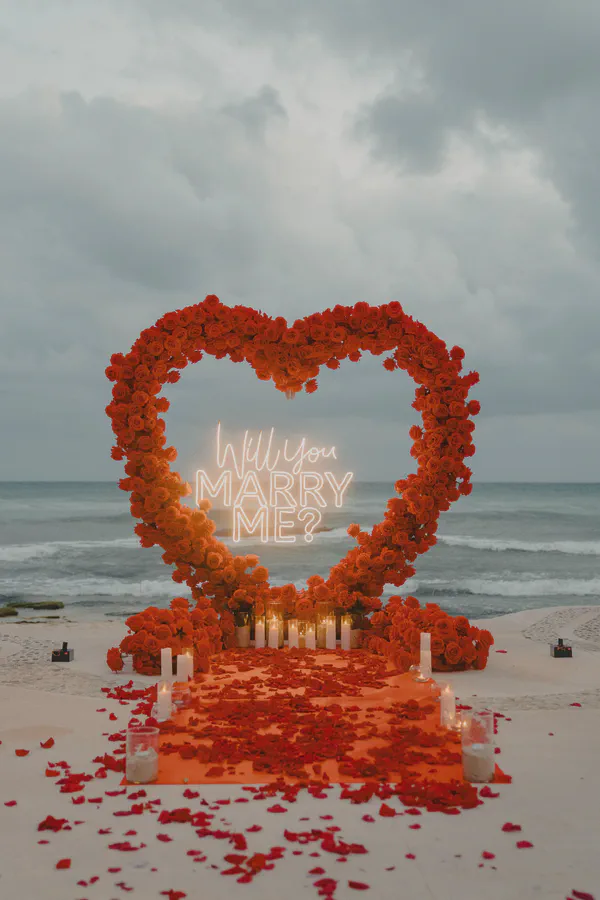 Propuesta en la playa con un imponente arco en forma de corazón, construido íntegramente con rosas rojas vibrantes que contrastan con un océano gris melancólico; un elegante letrero de neón y un camino de pétalos y portavelas de cristal sobre arena inmaculada crean un ambiente íntimo.