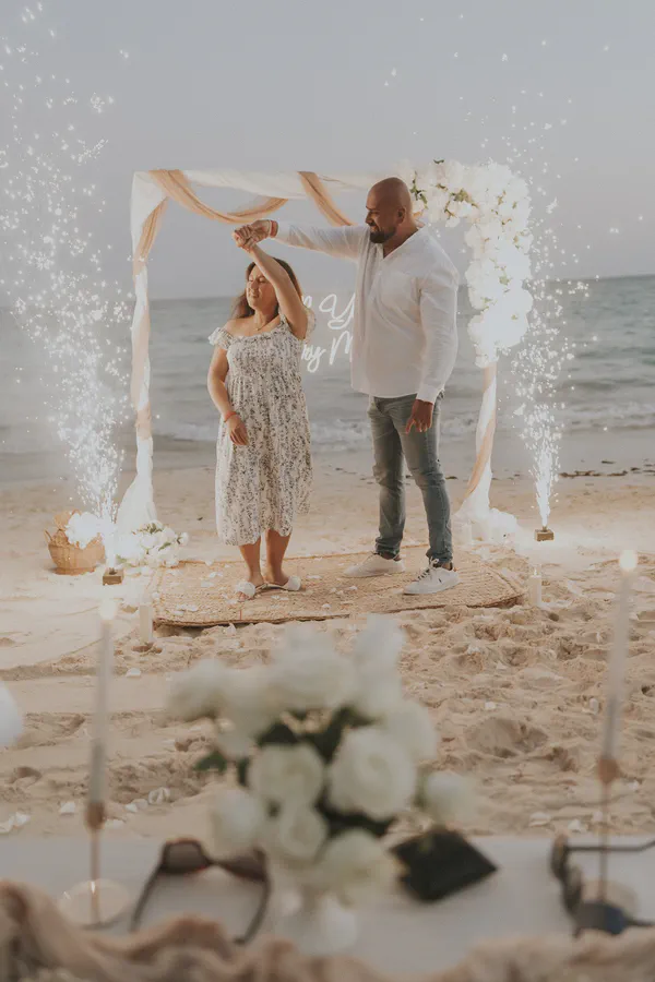 Montaje romántico en la paradisíaca Playa Akumal durante la hora dorada, con un elegante arco blanco decorado con drapeado rosado y arreglos florales; fuentes de chispas frías, velas en portavelas y rosas premium, complementados por una alfombra de yute natural, generan un ambiente etéreo y sofisticado.