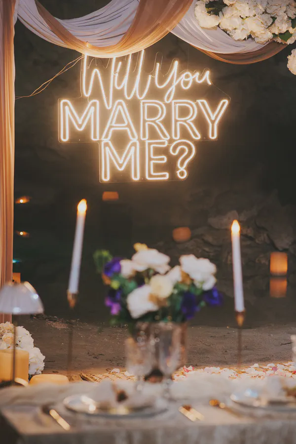 Impactante propuesta de matrimonio en una cueva íntima del Cenote Calavera, donde un letrero LED de neón 'Will You Marry Me?' se erige como pieza central; el drapeado de tela blanca, hortensias premium y velas en portavelas realzan un montaje de mesa refinado.