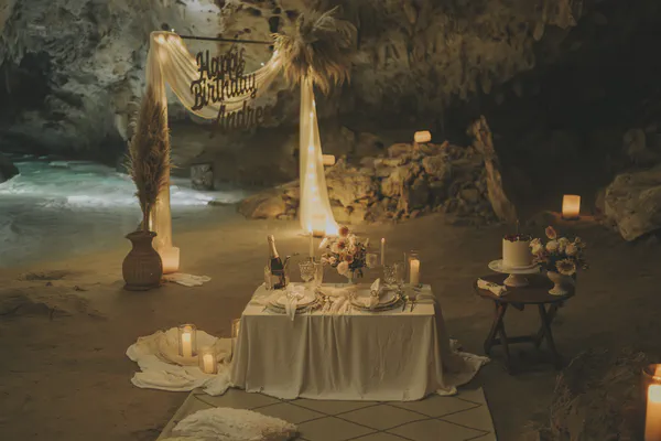 Cena privada de cumpleaños en un cenote natural cerca de Playa del Carmen, con una mesa elegantemente decorada para dos que se ilumina con cálida luz de velas; un banner personalizado se extiende sobre un arco de tela con pasto pampas seco, mientras cristalería fina crea ambiente lujoso.