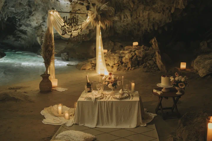 A candlelit table for two with champagne and a cake beside the water in a cave cenote, under a draped arch with a birthday sign.