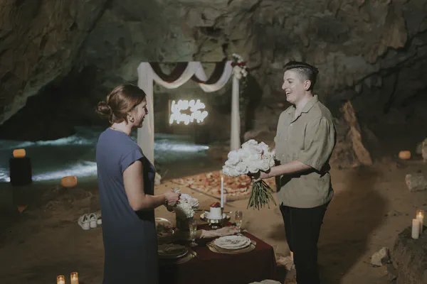 Cena romántica de lujo en un cenote místico, donde un montaje curado crea una atmósfera íntima entre paredes dramáticas iluminadas con luz de velas y un letrero de neón; una mesa vestida con vajilla fina se funde armoniosamente con el entorno rocoso.