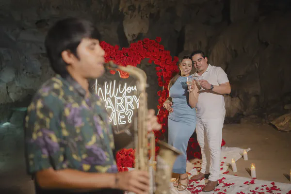 Propuesta de matrimonio en el Cenote Azul, donde paredes de piedra caliza iluminadas resaltan un letrero de neón 'Will You Marry Me?' junto a un elegante arco de rosas rojas; velas románticas y pétalos esparcidos se combinan con el toque musical de un saxofonista.
