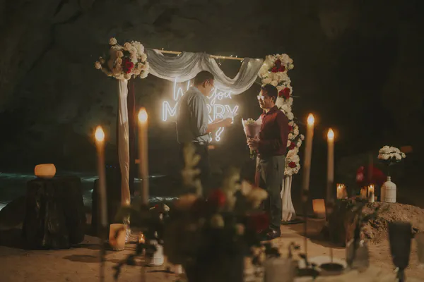 Propuesta en un cenote místico con un arco drapeado en blanco adornado con abundantes rosas blancas y rojas intensas; la iluminación estratégica, que incluye velas en portavelas y un moderno letrero de neón 'Will You Marry Me?', equilibra el brillo cálido y frío.