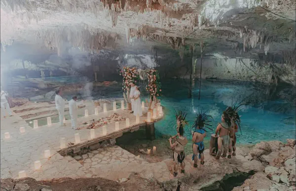 Ceremonia mística en el sagrado Cenote Suytun, con sus formaciones de piedra caliza y aguas turquesas cristalinas; un sendero de luminarias conduce a un elegante arco floral decorado con flores coral y melocotón, mientras performers mayas en atuendos ceremoniales aportan autenticidad.