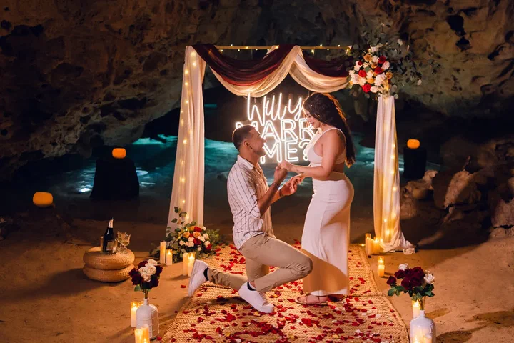 Hombre arrodillado pidiendo matrimonio bajo un arco de telas con letrero de neón “Will You Marry Me?” en un cenote