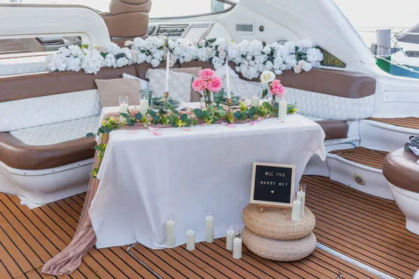 Propuesta en yate de lujo navegando frente a Playa Norte de Isla Mujeres, con un interior exquisitamente decorado que combina asientos de cuero acolchado blanco, guirnaldas de rosas y hortensias, y una sofisticada disposición de comedor con manteles y acentos en dorado.