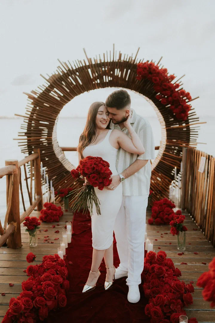 Pareja abrazada bajo un arco de carrizo en forma de sol con rosas rojas en un muelle junto al mar