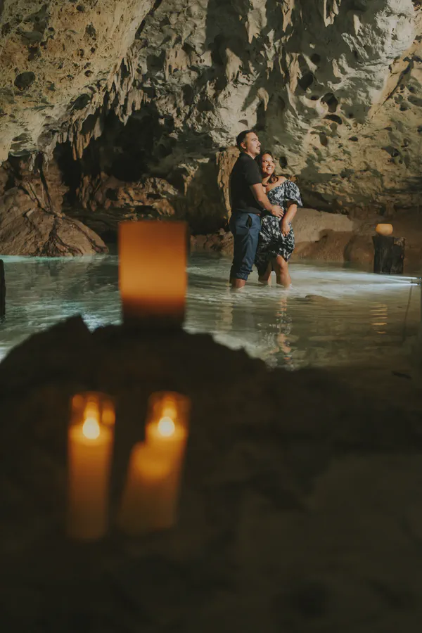 Propuesta privada en el Cenote Jardín del Edén, donde aguas cristalinas y formaciones milenarias de piedra caliza se iluminan con cálida luz de velas; estalactitas y reflejos ámbar en el agua enmarcan a la pareja en un ambiente íntimo, ideal para celebraciones exclusivas.