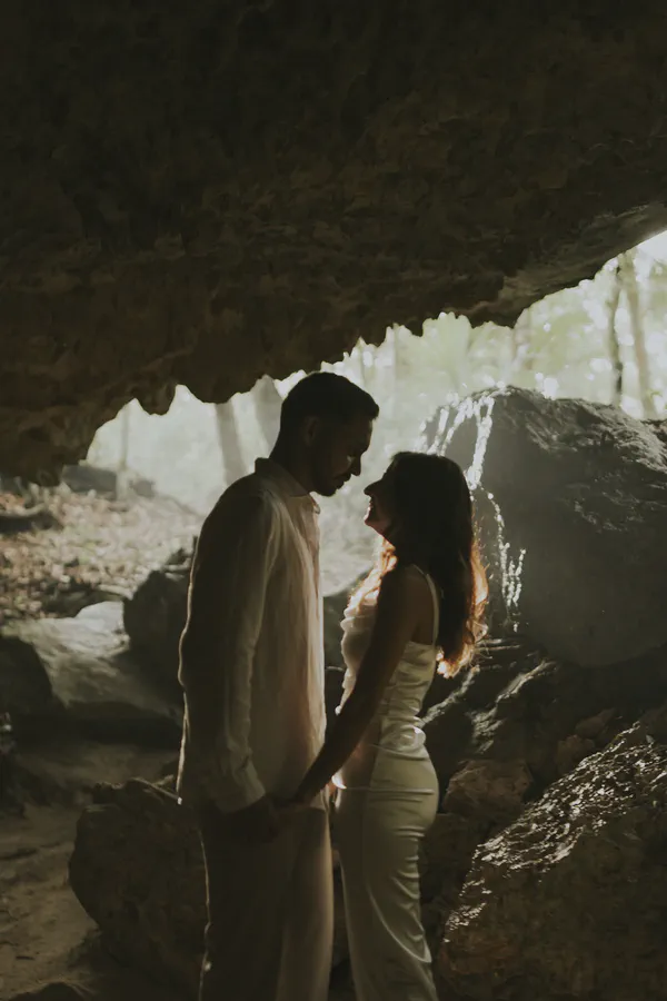 Momento romántico capturado en Cenote Casa Tortuga, donde la silueta de una pareja elegantemente vestida se destaca contra la luz etérea filtrada a través de la abertura natural de la caverna; las texturas de piedra caliza y la iluminación cinematográfica evocan intimidad.