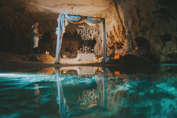 Propuesta en caverna de cenote con un asombroso reflejo en aguas turquesas; un arco drapeado en blanco con toques en azul y acentos florales, junto a un letrero de neón, y una mesa decorada con arreglos púrpuras y velas, conforman la fusión ideal de elementos naturales y de lujo.