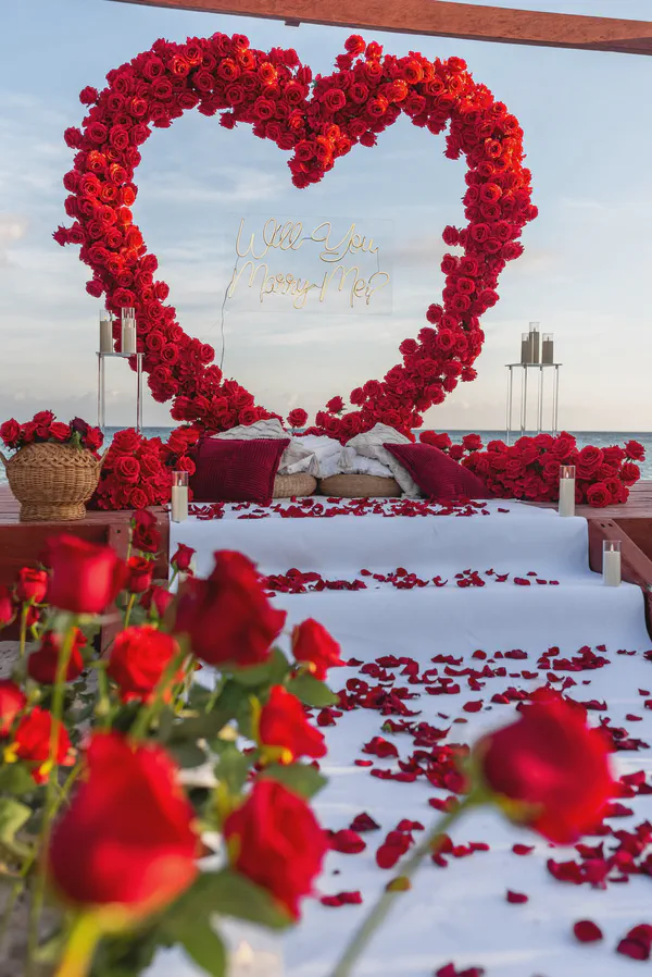 Propuesta frente al mar en la zona hotelera de Cancún con un dramático arco en forma de corazón, construido enteramente con rosas rojas vibrantes y un letrero dorado central 'Will You Marry Me?', sobre una plataforma elevada con escalones y pétalos esparcidos.