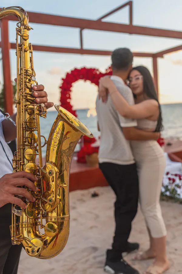 Propuesta romántica junto al mar durante la hora dorada en la zona norte de Playa del Carmen, destacada por un impresionante saxofón dorado en primer plano y una estructura con marco rojo que enmarca un arco de rosas rojas; la iluminación suave del atardecer resalta el brillo del latón.