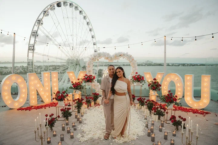 Una pareja en una azotea entre letras luminosas “ONLY YOU”, rosas rojas y velas, con una rueda de la fortuna detrás