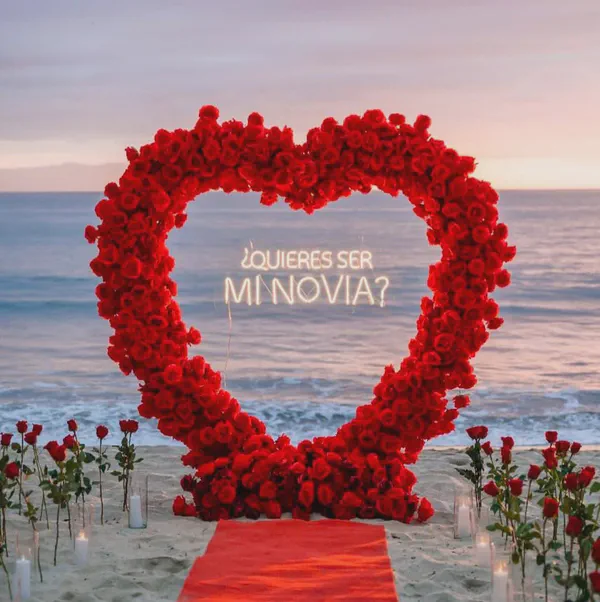 Propuesta en la exclusiva Playa Maroma con un majestuoso arco en forma de corazón compuesto de rosas rojas vibrantes; el arco enmarca un letrero de neón en español '¿QUIERES SER MI NOVIA?' iluminado en blanco cálido, mientras una alfombra roja crea un camino romántico.