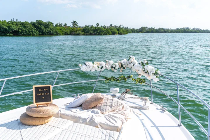 Proa de un yate con cojines, una guirnalda de rosas blancas en el barandal y una pizarra con “Will you marry me?”, en una laguna rodeada de manglar