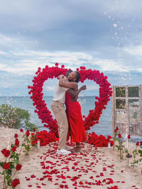 Una pareja alegre se abraza apasionadamente durante una romántica propuesta en la playa. La mujer, vistiendo un vestido rojo impresionante y tacones plateados, está envuelta en un abrazo sincero por su pareja, quien está vestido con una camisa blanca y pantalones beige. Se encuentran ante un impresionante arco en forma de corazón compuesto completamente de rosas rojas vibrantes, creando un telón de fondo hipnotizante contra una vista serena del océano bajo un cielo suave y nublado. Fuegos artificiales chispeantes añaden un toque de magia a la escena, bañando el momento con luces celebratorias. Pétalos de rosa están esparcidos en la arena creando un sendero que lleva a la pareja, realzando la atmósfera romántica y celebratoria. La escena general está llena de amor, felicidad y emoción, capturada en una imagen impresionante rica en color y emoción. El letrero "Will you marry me?" añade un toque final al momento perfecto de la propuesta.