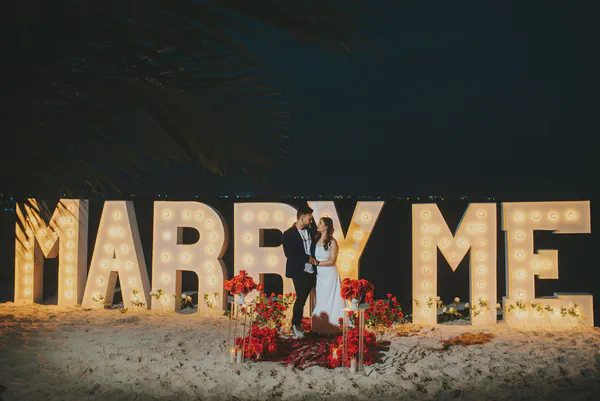 Una romántica propuesta en la playa por la noche. Una pareja, un hombre con un blazer azul oscuro y una mujer con un vestido blanco, se encuentran muy cerca mirándose a los ojos frente a grandes letras iluminadas "Marry Me". Las letras son blancas con bombillas cálidas y brillantes delineando cada letra. La escena está ambientada en una playa arenosa, con el océano oscuro y un paisaje urbano débilmente visible en el fondo. Rodeando a la pareja hay elegantes arreglos de rosas rojas profundas y velas pilares, añadiendo al ambiente romántico. El ambiente general es alegre, íntimo y celebratorio, capturando un momento especial de compromiso y amor. La iluminación cálida contrasta bellamente con el cielo nocturno fresco, realzando la atmósfera mágica de la propuesta en la playa.