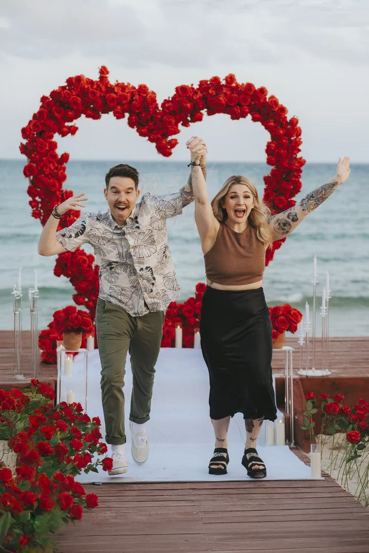 Pareja celebrando frente a un corazón de rosas rojas al final de un pasillo en la playa