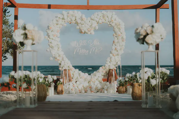 Una escena romántica de propuesta de boda en la playa está montada bajo una pérgola de madera. Un gran arreglo floral en forma de corazón, predominantemente rosas blancas y blanquecinas, sirve como telón de fondo. Un letrero de neón dentro del corazón dice "Will You Marry Me?" en una escritura delicada. Arreglos florales blancos más pequeños y velas en contenedores de vidrio y tejidos flanquean el corazón, añadiendo al ambiente elegante e íntimo. El océano es visible en el fondo, añadiendo una sensación serena y pacífica a la escena. La paleta de colores general es suave, con las flores blancas contrastando bellamente contra la madera marrón cálida y el océano azul. La escena evoca sentimientos de amor, esperanza y expectativa.